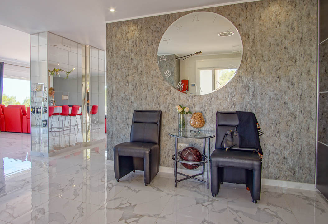 Zona de recibidor con suelos de mármol pulido, pared de piedra texturizada y un gran espejo circular decorativo. Incluye mobiliario de cuero y columnas revestidas de espejos.