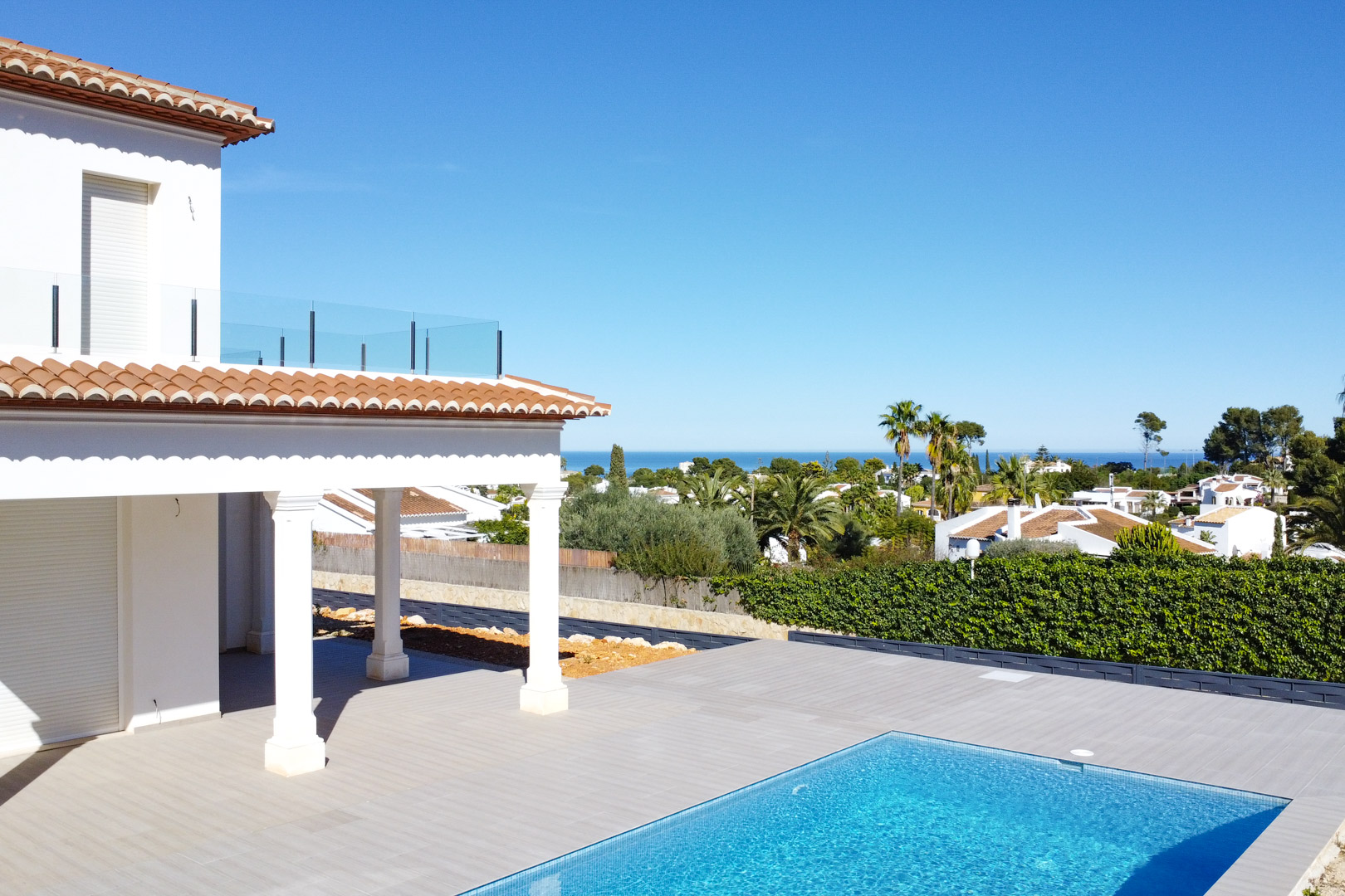 Vista exterior de la villa con terraza de gres porcelánico, piscina de mosaico azul y porche con columnas. Al fondo se aprecia el mar Mediterráneo y el entorno de Pinosol.