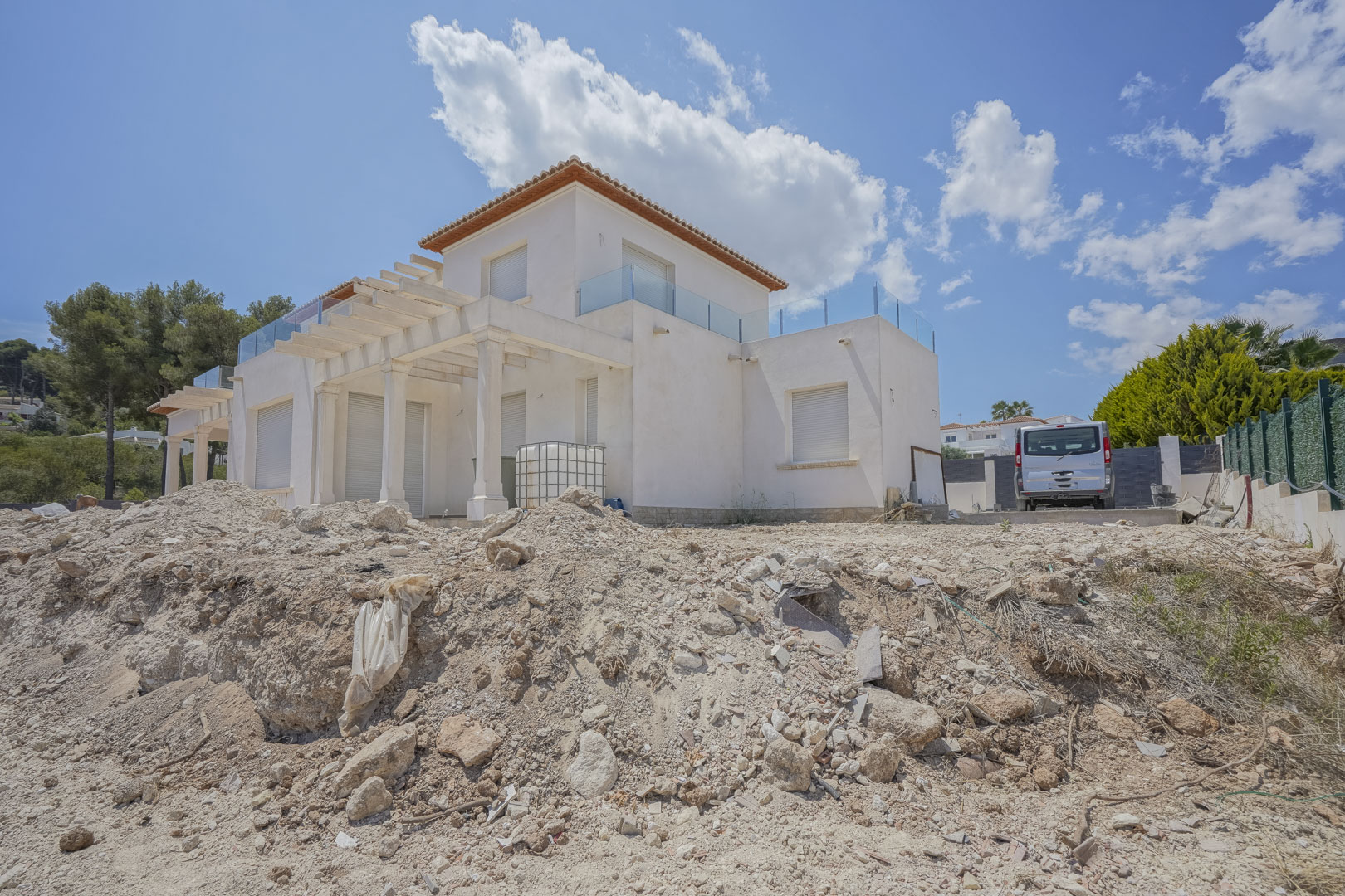 Vista exterior de la villa en fase final de obra, mostrando la fachada blanca, pérgolas con columnas y terrazas superiores con barandillas de cristal.