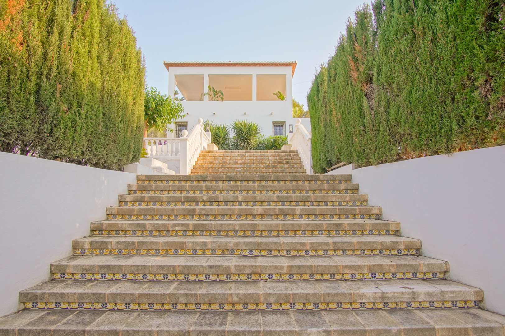 Amplia escalinata de piedra con contrahuellas de azulejo decorativo, flanqueada por altos cipreses que conducen a la fachada blanca y terraza superior de la villa.