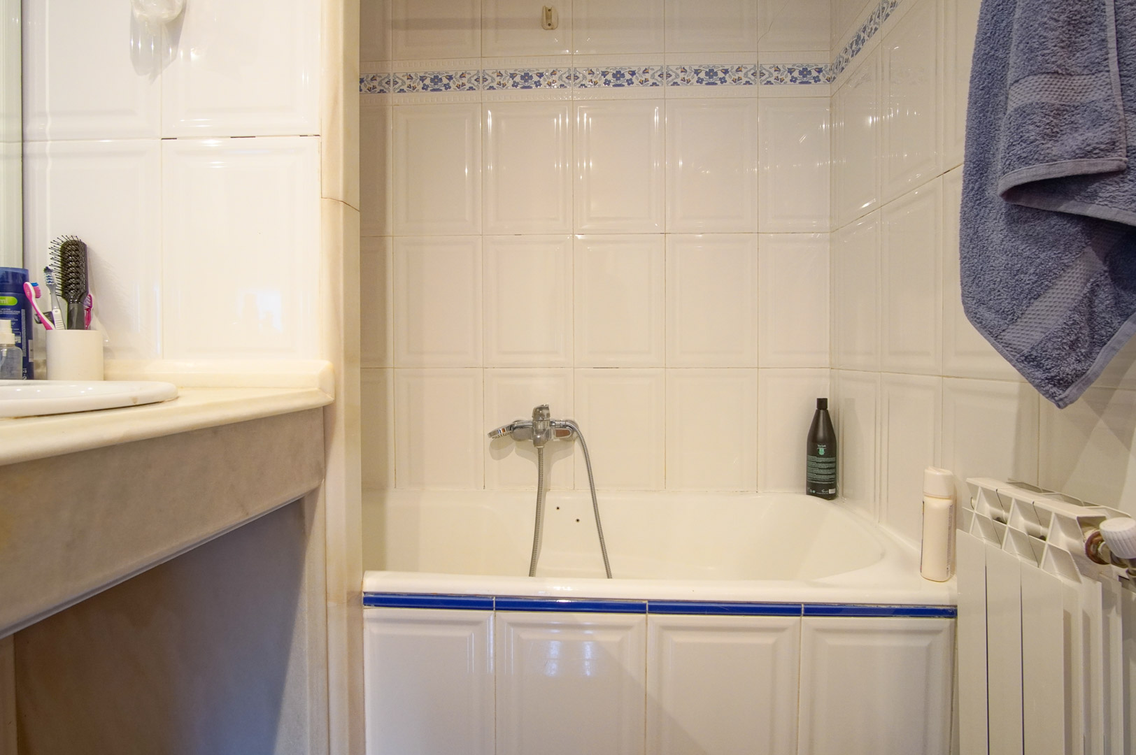 Baño funcional con bañera integrada, paredes de azulejo blanco con detalles florales azules y encimera de mármol. Dispone de radiador y grifería cromada clásica.