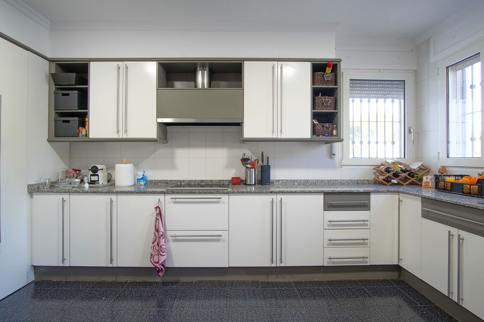 Cocina independiente con armarios blancos, encimera de granito gris y placa de inducción. Dispone de amplios ventanales y suelo de baldosa oscura moteada.