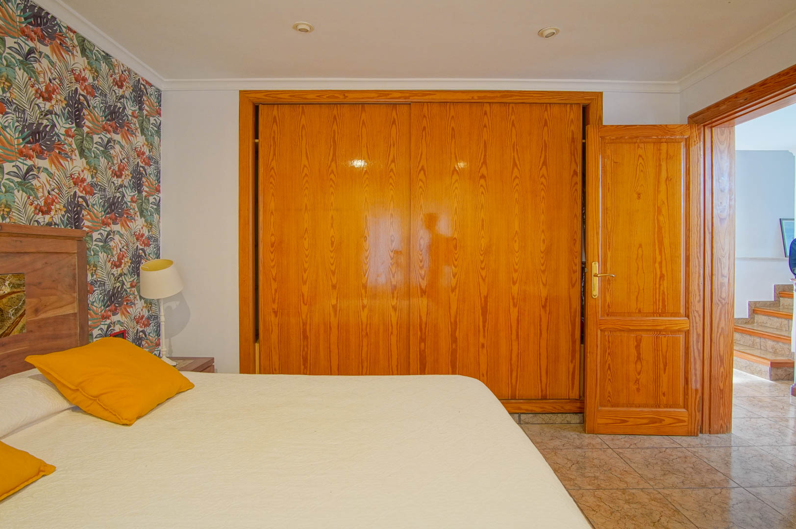 Dormitorio con amplio armario empotrado de madera, pared de acento con papel tapiz tropical y suelos cerámicos. La puerta abierta muestra el acceso a la escalera.