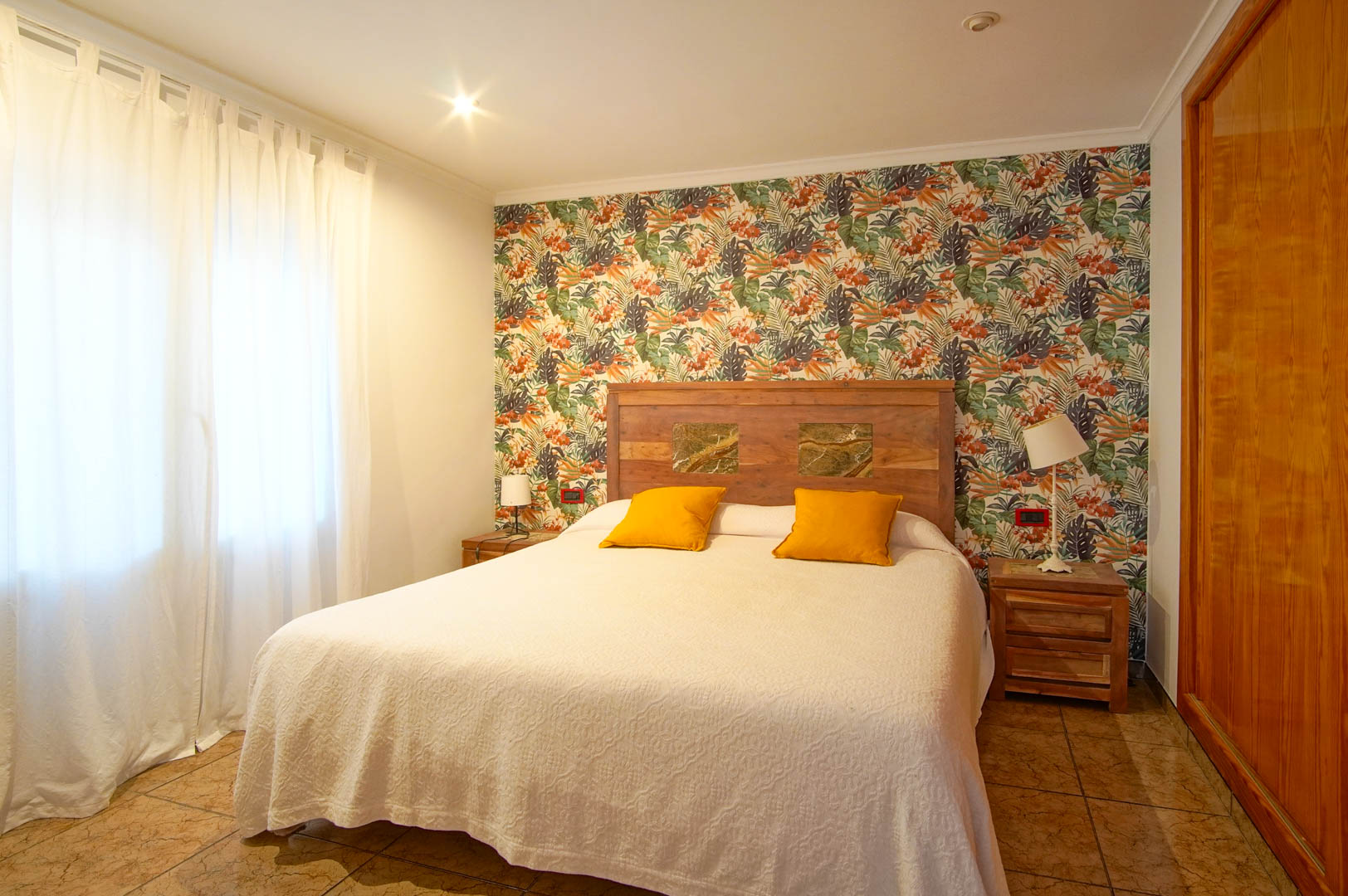 Dormitorio con papel pintado de motivos tropicales, cabecero de madera con incrustaciones de piedra y armario empotrado lateral.