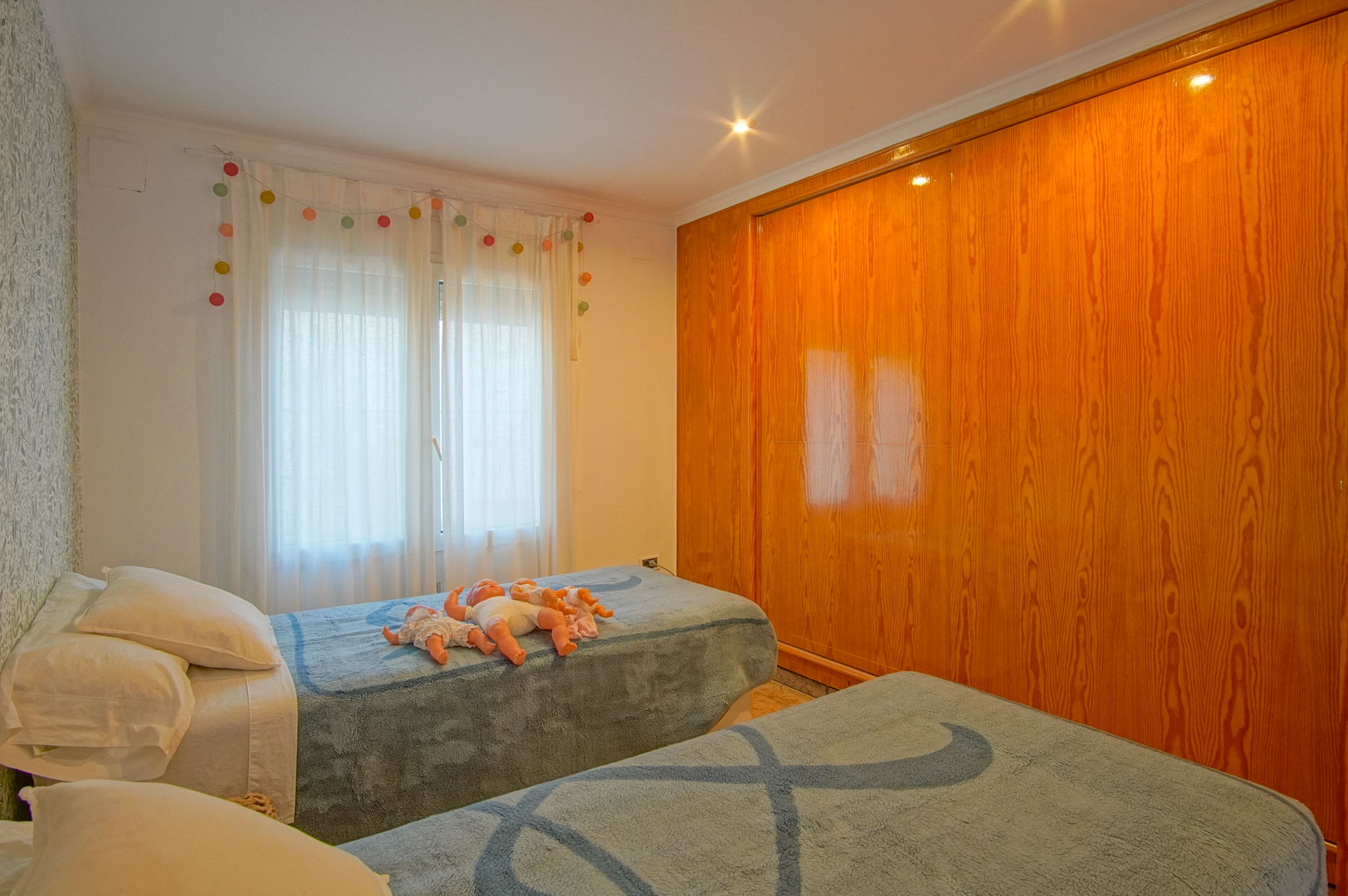 Dormitorio luminoso con dos camas individuales, papel pintado floral y un amplio armario empotrado de madera natural con acabado brillante.