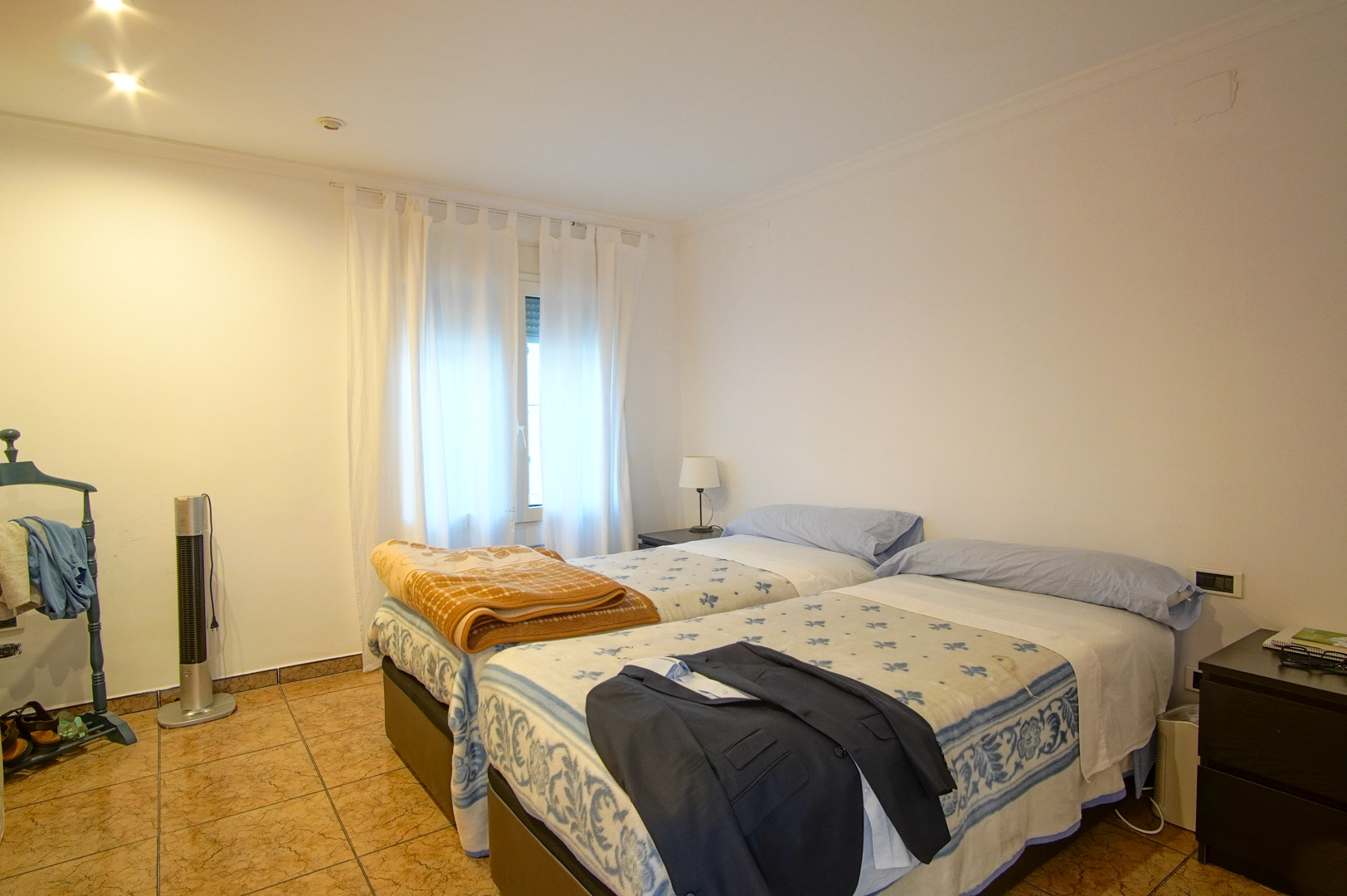 Dormitorio luminoso con dos camas individuales, suelos de gres cerámico y paredes blancas. Incluye iluminación empotrada y luz natural a través de una ventana con cortinas.