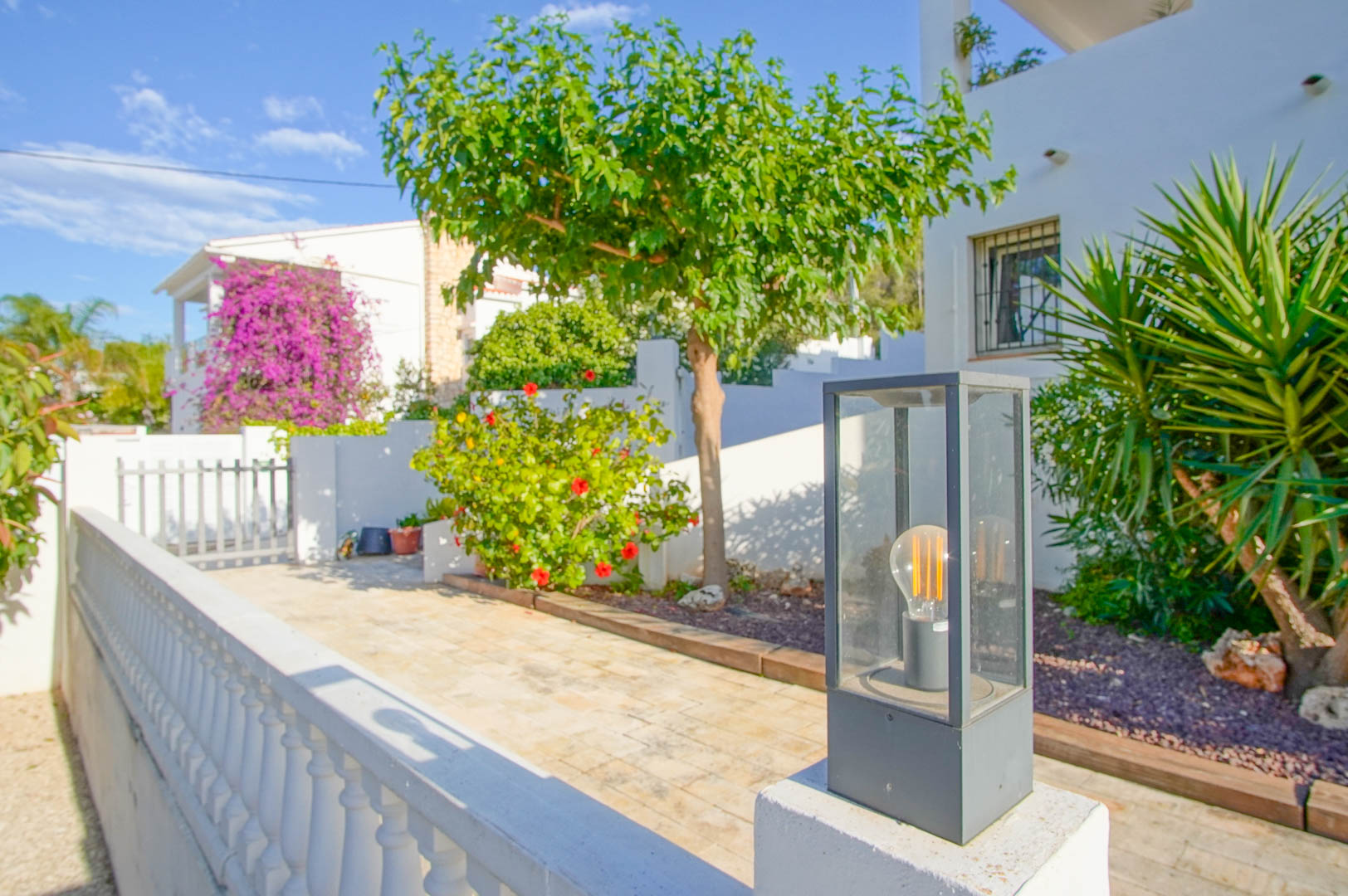 Entrada exterior con pavimentación de piedra, iluminación moderna de diseño y vegetación mediterránea que incluye hibiscos y buganvillas sobre fachadas blancas.