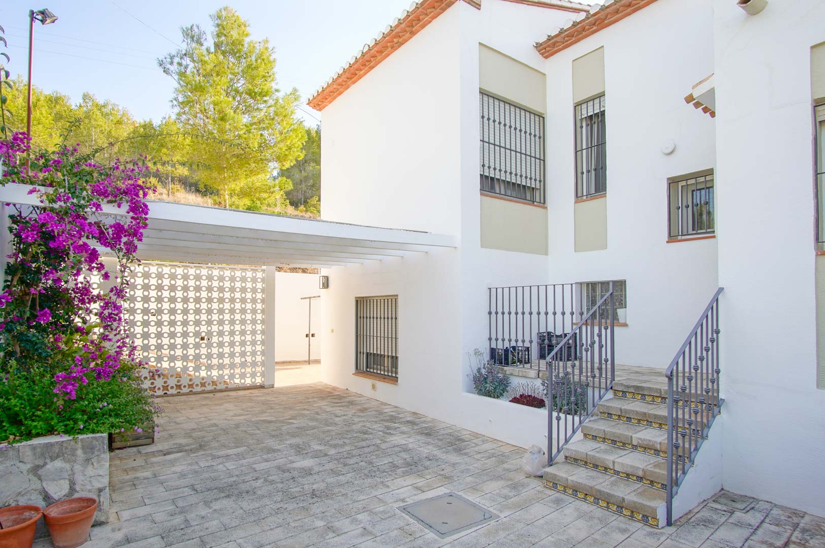 Entrada exterior con pavimentación de piedra, zona de aparcamiento con pérgola blanca y escaleras de acceso con azulejos decorativos y barandilla de forja.