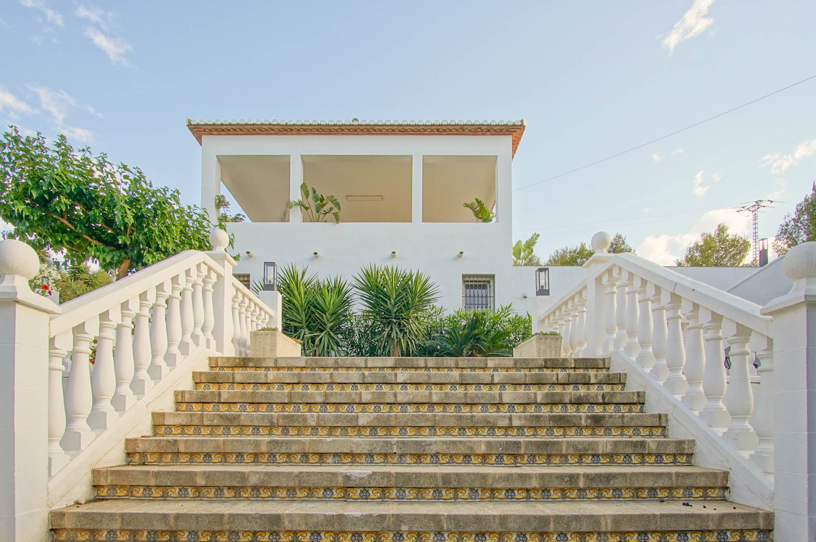 Escalera exterior de piedra con contrahuellas de cerámica decorativa y balaustrada blanca, que conduce a la terraza superior y fachada de la villa.