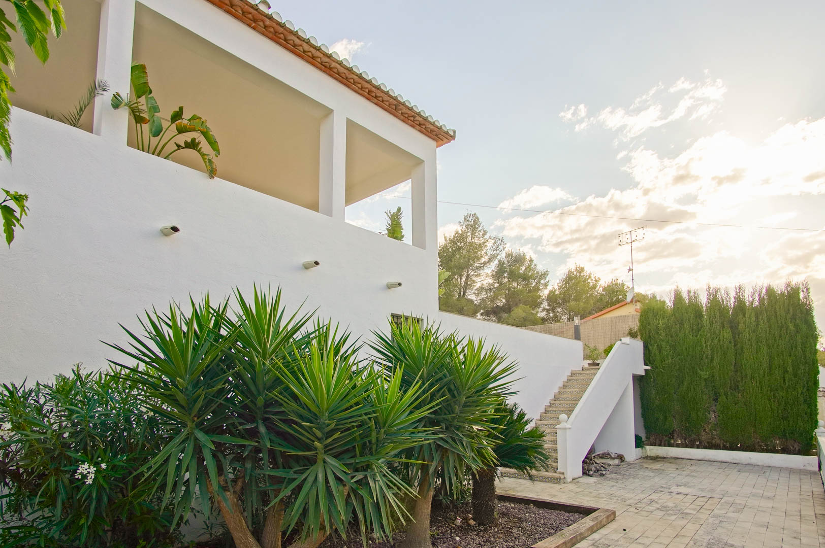 Fachada blanca de estilo mediterráneo con terraza cubierta y pilares cuadrados. El exterior incluye una escalera con azulejos decorativos y un jardín con yucas y cipreses.