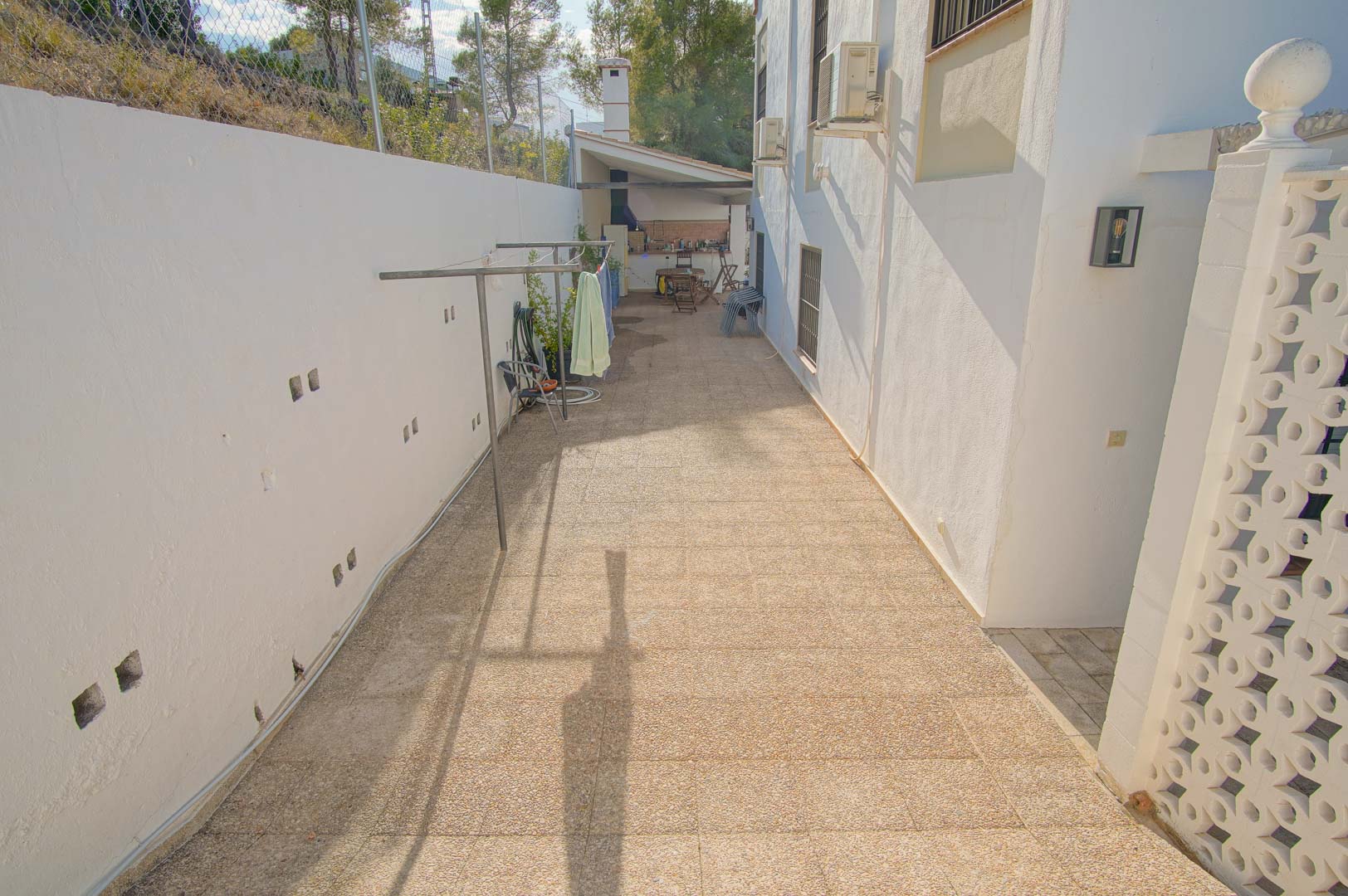 Pasillo lateral pavimentado con zona de tendedero, que conduce directamente al área de barbacoa cubierta y la cocina exterior.