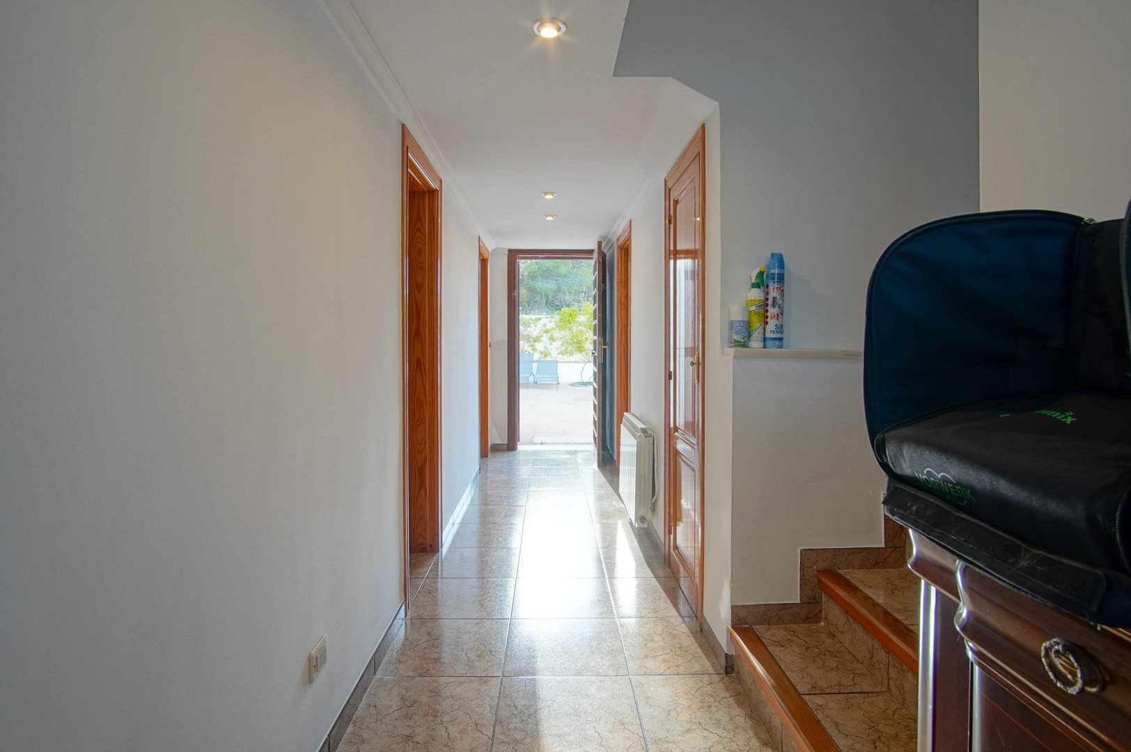 Pasillo luminoso con suelos cerámicos, marcos de madera y focos empotrados que conduce a una terraza exterior. Incluye acceso a las escaleras laterales.