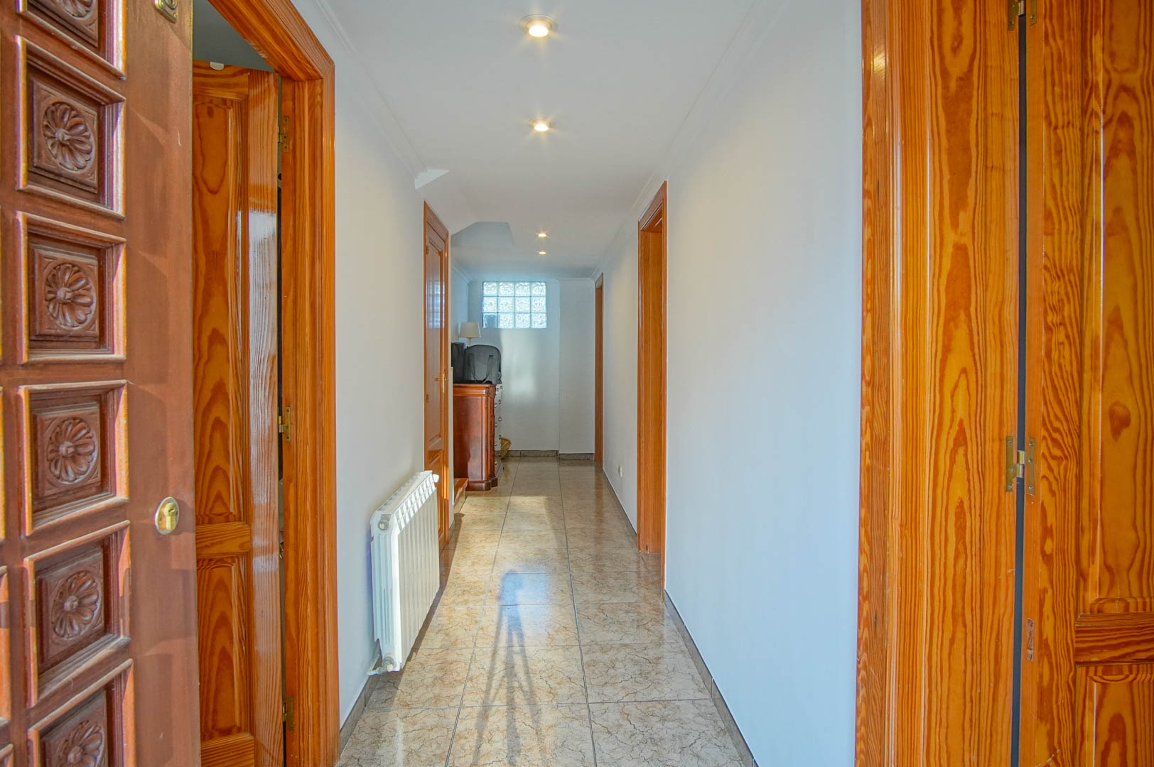 Pasillo luminoso con suelos cerámicos, puertas de madera tallada y calefacción central. Incluye iluminación empotrada y una ventana de bloques de vidrio al fondo.