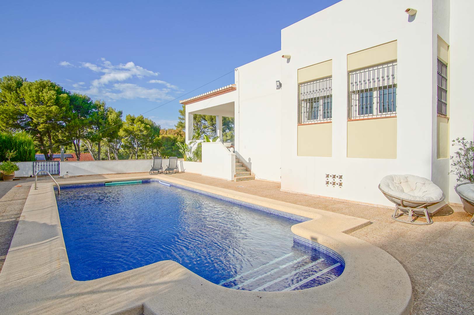 Piscina privada con escaleras romanas y terraza de piedra, situada junto a la fachada blanca de la villa con vistas a la zona de pinos de Las Rotas.