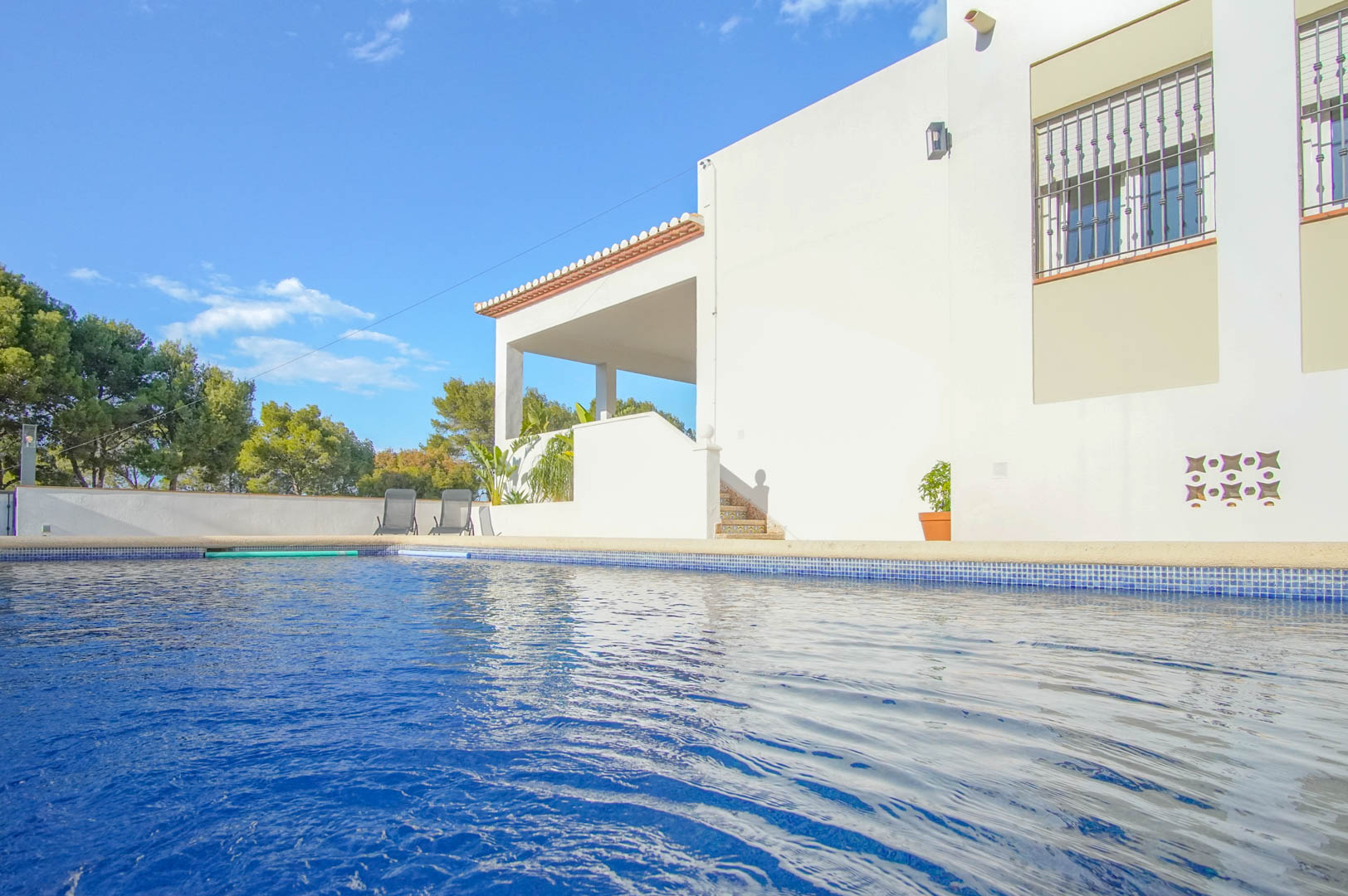 Piscina privada con gresite azul junto a la fachada blanca del chalet, que cuenta con una terraza cubierta y acceso mediante escaleras cerámicas en un entorno arbolado.