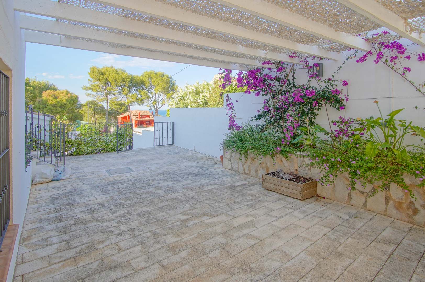 Terraza exterior pavimentada con pérgola blanca y malla de sombra, decorada con jardineras de piedra y buganvillas, ofreciendo vistas despejadas al entorno natural y al mar.