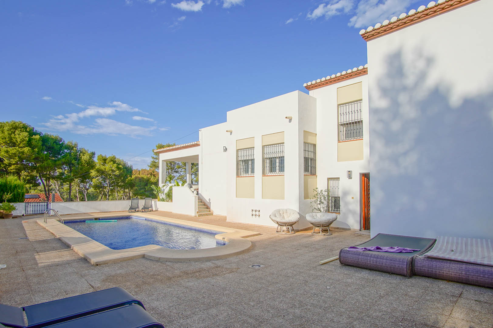 Vista exterior de la villa con fachada blanca, destacando la piscina privada y la amplia terraza pavimentada rodeada de vegetación mediterránea y pinos.