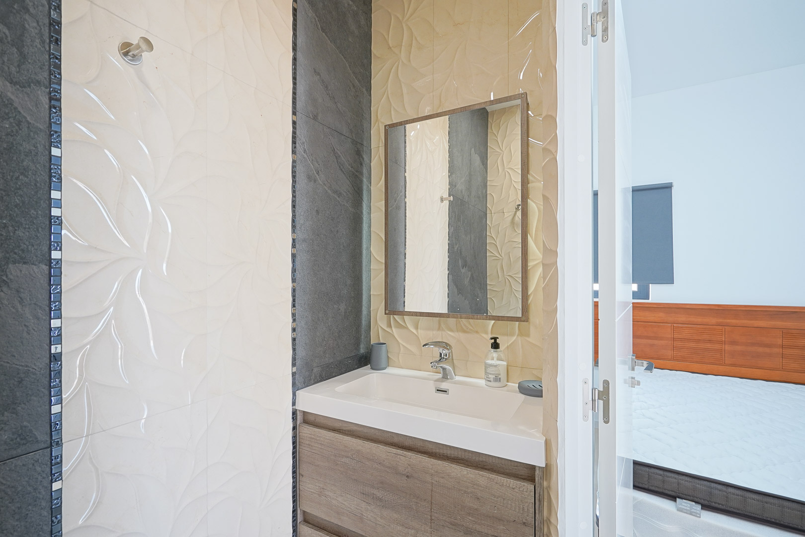 Baño ensuite con lavabo integrado, mueble de madera y paredes decoradas con azulejos texturizados en relieve y detalles en gris oscuro.