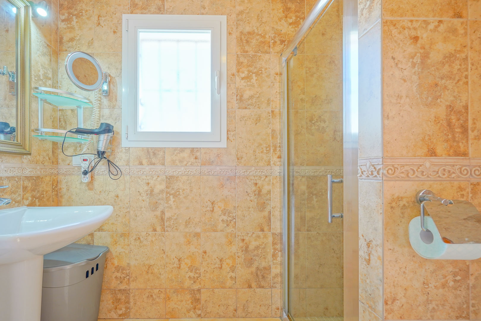 Cuarto de baño con azulejos en tonos tierra, lavabo de pedestal blanco y ducha con mampara de cristal. Incluye ventana oscilobatiente y accesorios cromados.