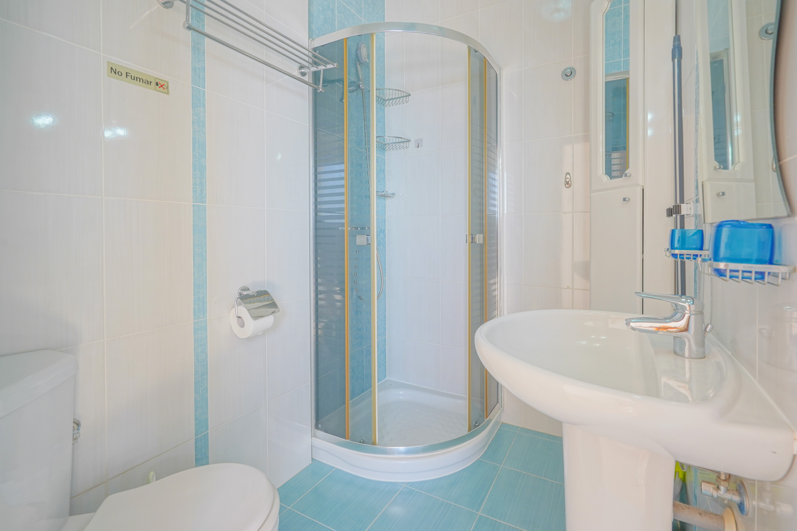 Cuarto de baño funcional con azulejos en blanco y azul, equipado con una cabina de ducha de rincón curva y lavabo de pedestal blanco.