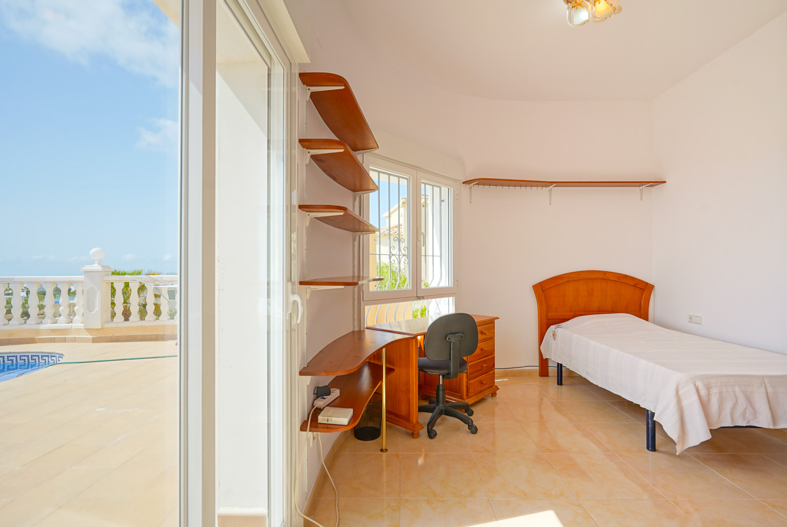 Dormitorio con cama individual, escritorio de madera y estanterías integradas. Acceso directo a la terraza de la piscina mediante amplias puertas correderas.