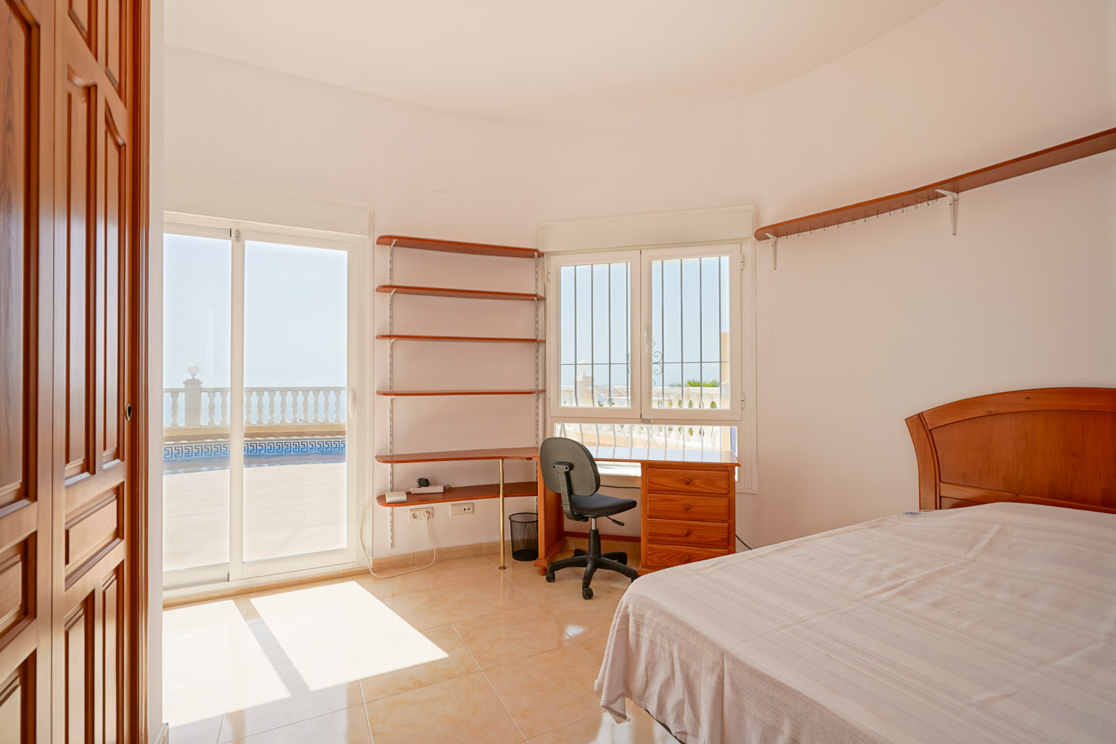 Dormitorio con zona de trabajo, estanterías de madera y acceso directo a la terraza mediante puertas correderas. Suelos de gres y abundante iluminación natural.