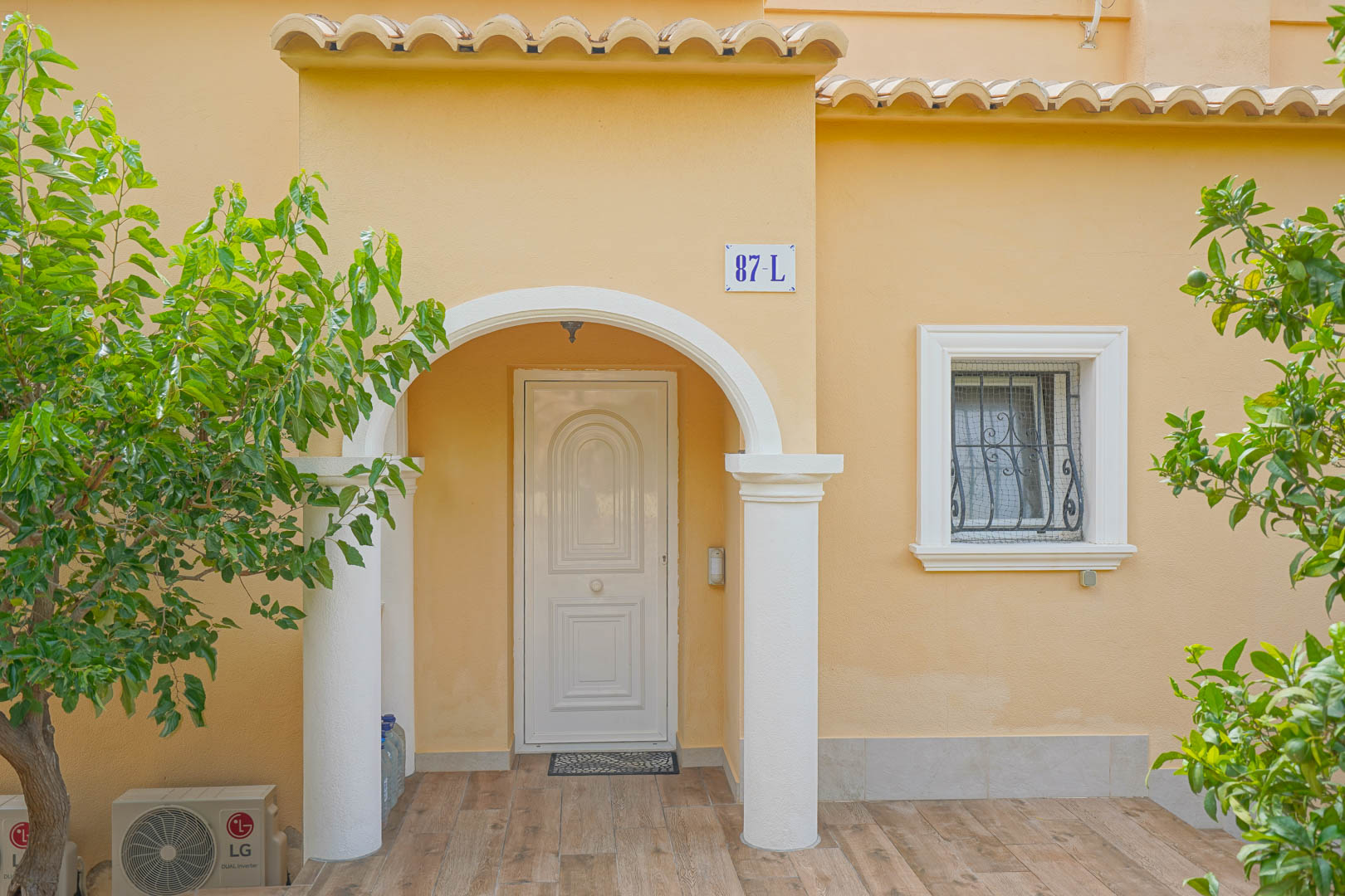 Entrada principal con porche de arco blanco y columnas, fachada en tono ocre y suelos cerámicos de efecto madera rodeados de vegetación mediterránea.
