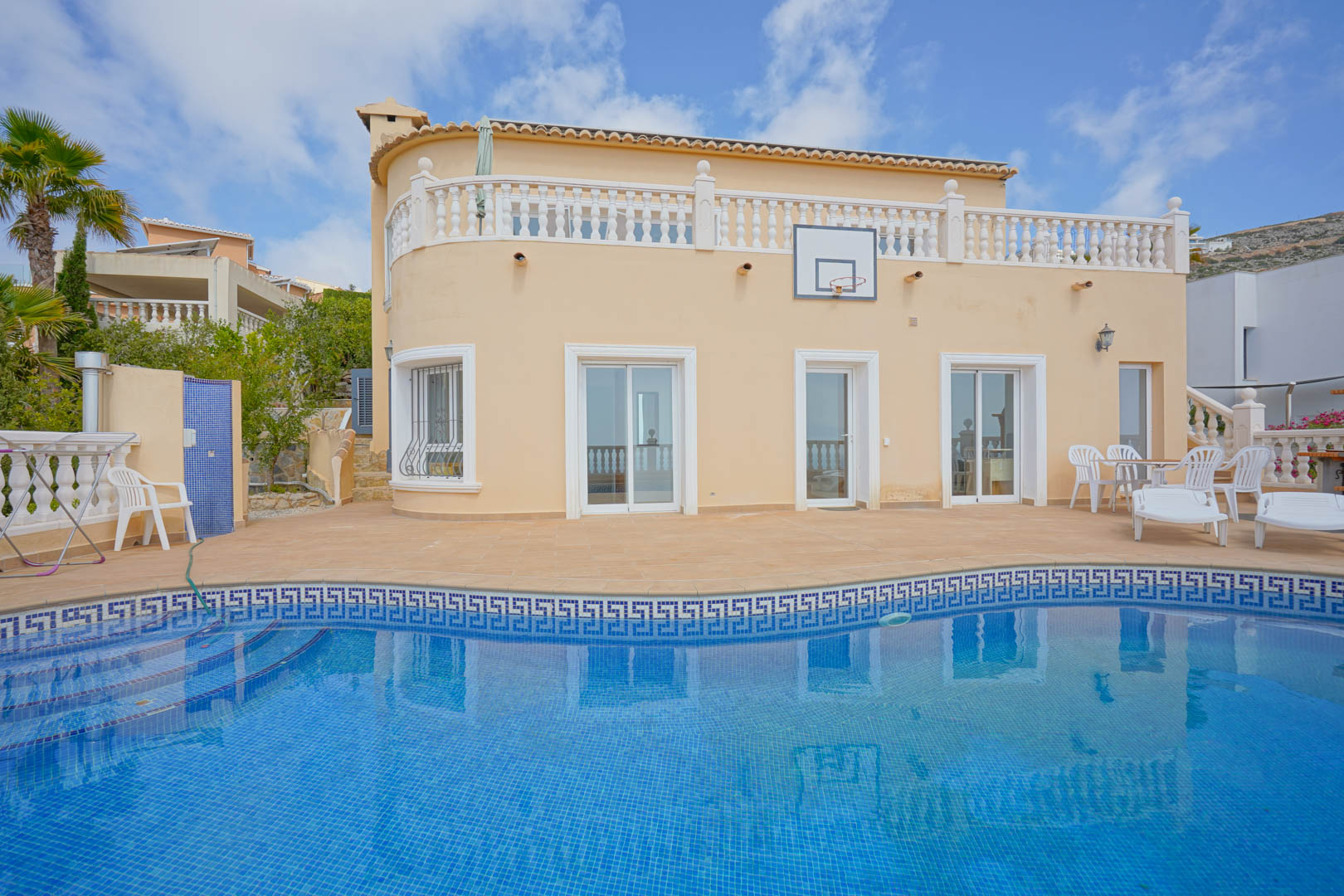 Fachada curva de la villa con piscina de mosaico azul, amplia terraza de terracota, zona de estar exterior y balaustradas blancas bajo un cielo despejado.