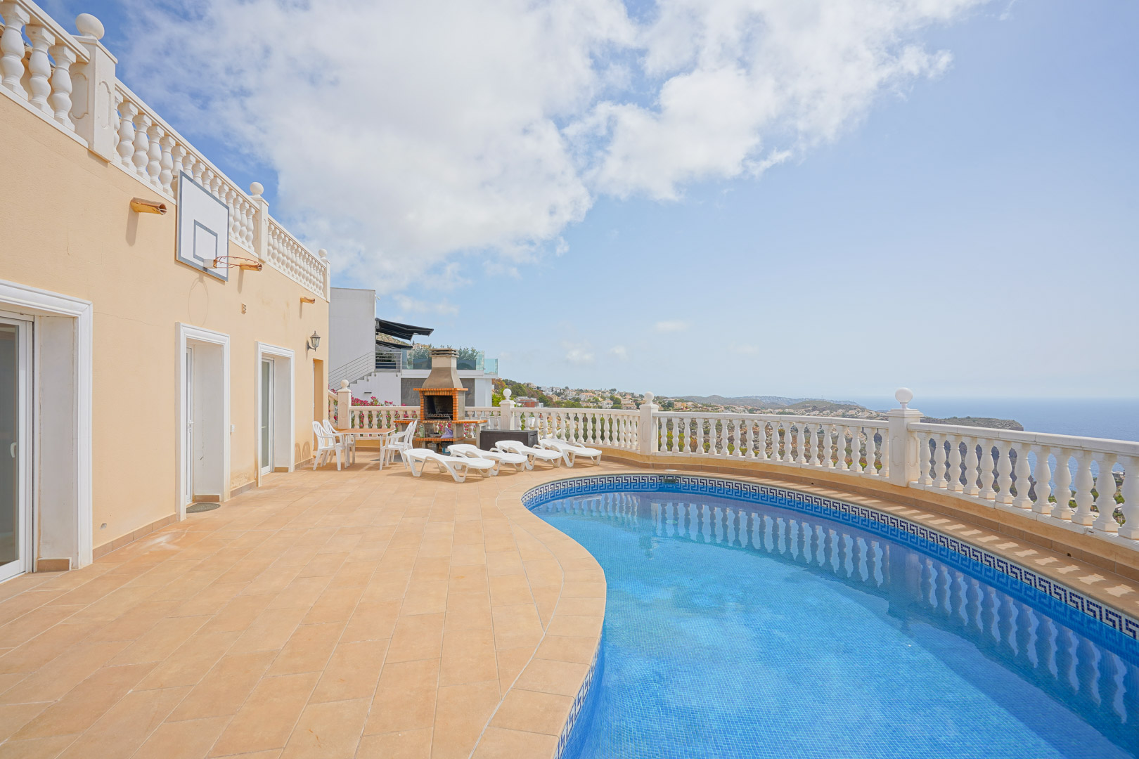 Terraza exterior con piscina de forma irregular, zona de barbacoa de obra y vistas panorámicas al mar Mediterráneo bajo una balaustrada blanca clásica.