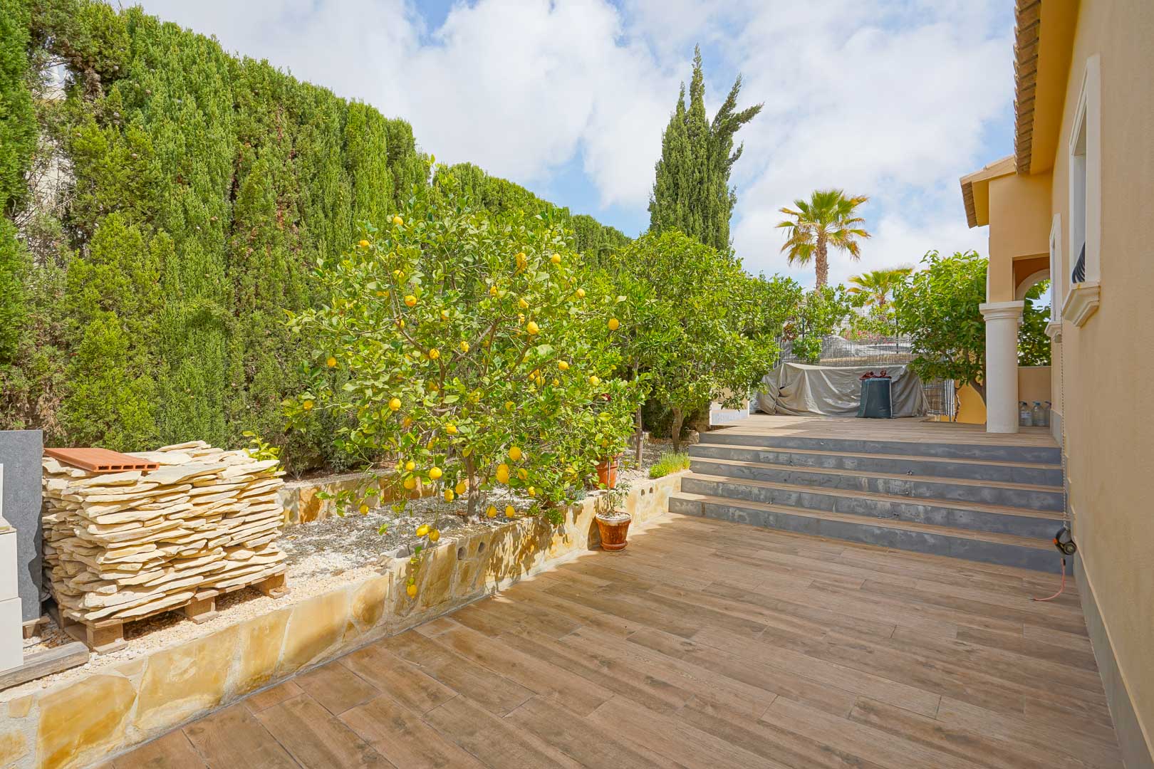 Terraza exterior con suelo cerámico de imitación madera, rodeada de limoneros y setos de ciprés, con escalones de piedra que conectan los niveles del jardín.