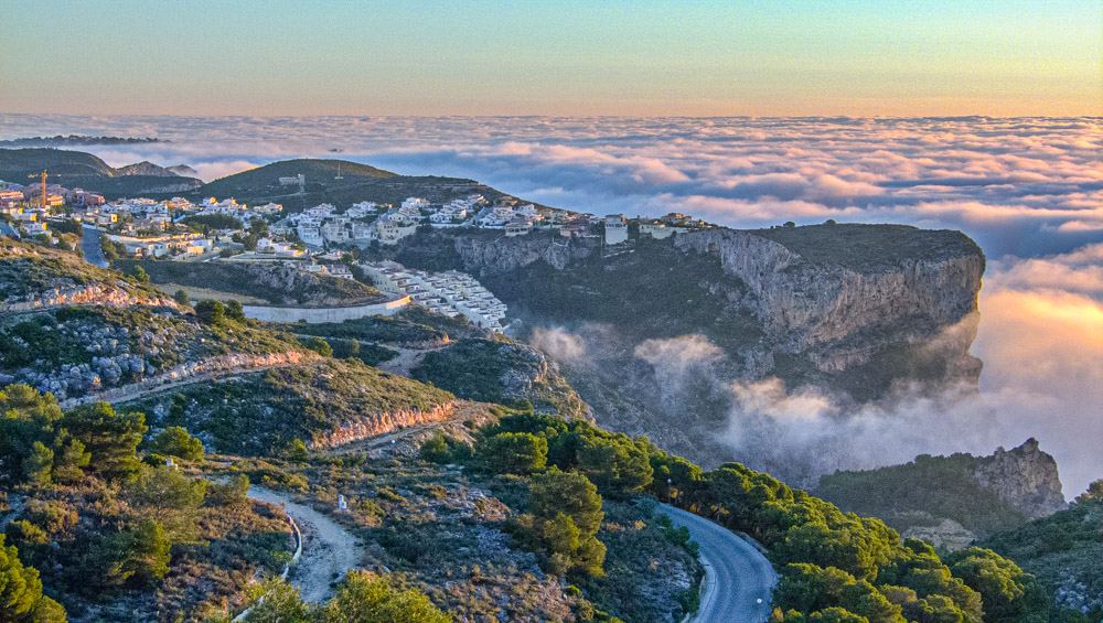 Vista panorámica de los acantilados de Cumbre del Sol sobre un mar de nubes al atardecer, destacando la orografía costera y las zonas residenciales integradas.