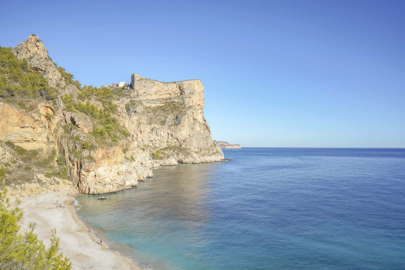 Vista panorámica de los acantilados de Cumbre del Sol y la playa de Cala Moraig, bañados por las aguas cristalinas del mar Mediterráneo bajo un cielo despejado.