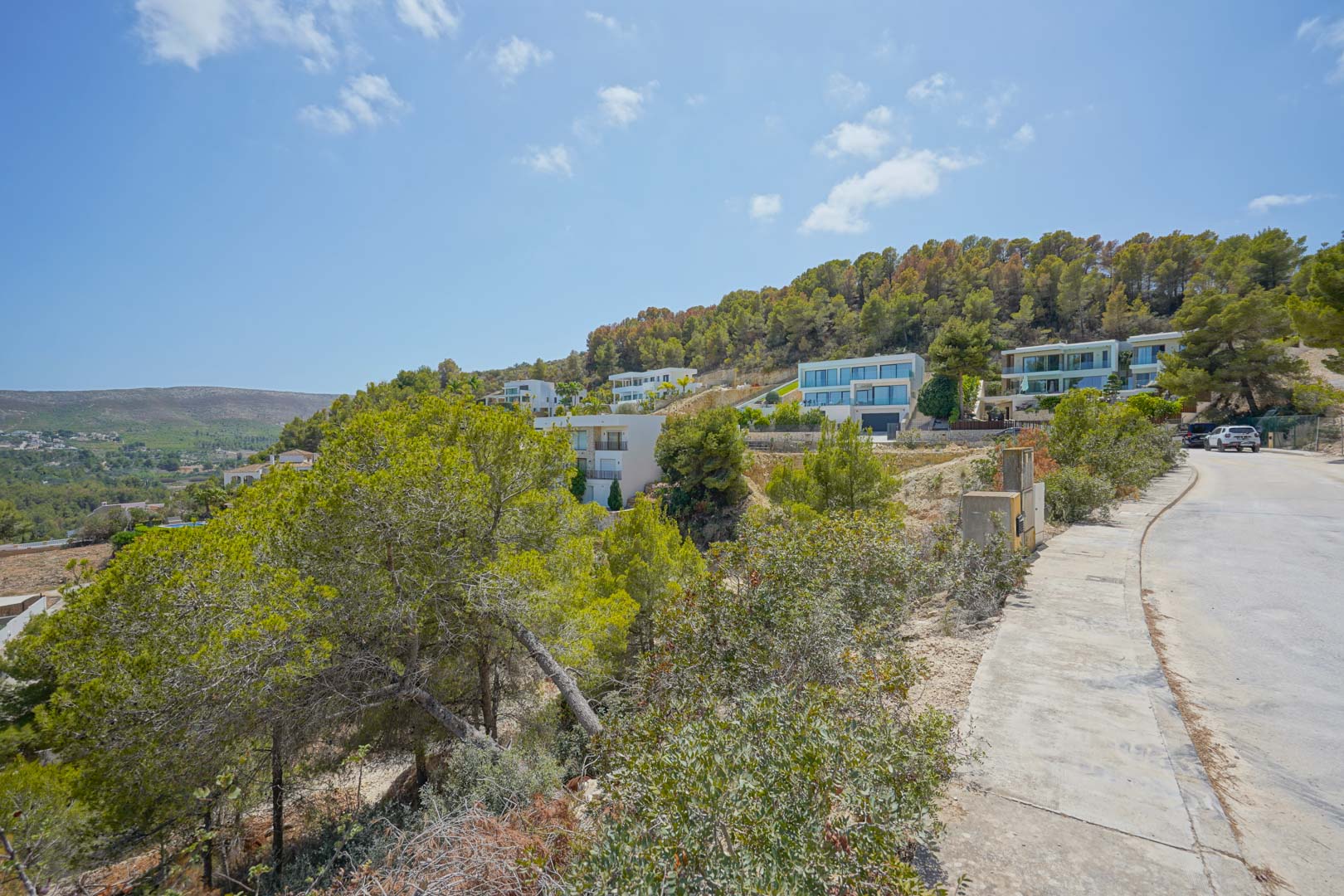 Vista de la infraestructura urbana con aceras y conexiones eléctricas, rodeada de pinos y villas modernas en una ladera con pendiente.