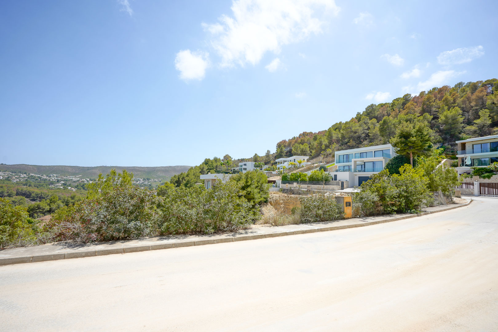 Vista desde la calle asfaltada de la urbanización, mostrando la infraestructura moderna y villas de diseño contemporáneo integradas en la ladera boscosa.