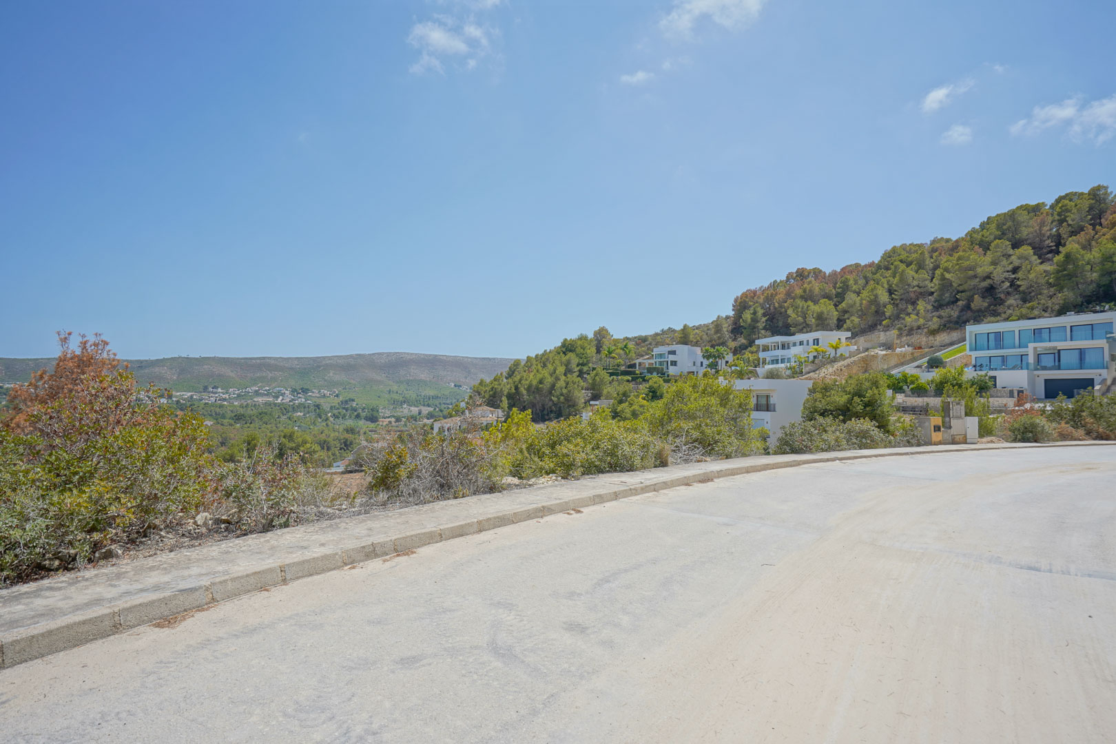 Vista desde la calle pavimentada de la urbanización, mostrando la topografía inclinada y las villas modernas integradas en la colina con vistas despejadas al valle.