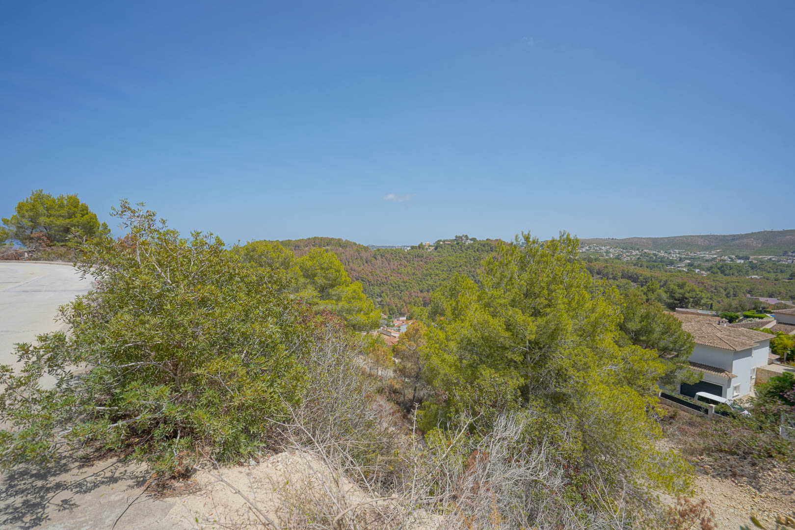 Vista elevada desde la parcela que muestra el acceso pavimentado y las colinas circundantes con vegetación mediterránea bajo un cielo despejado.