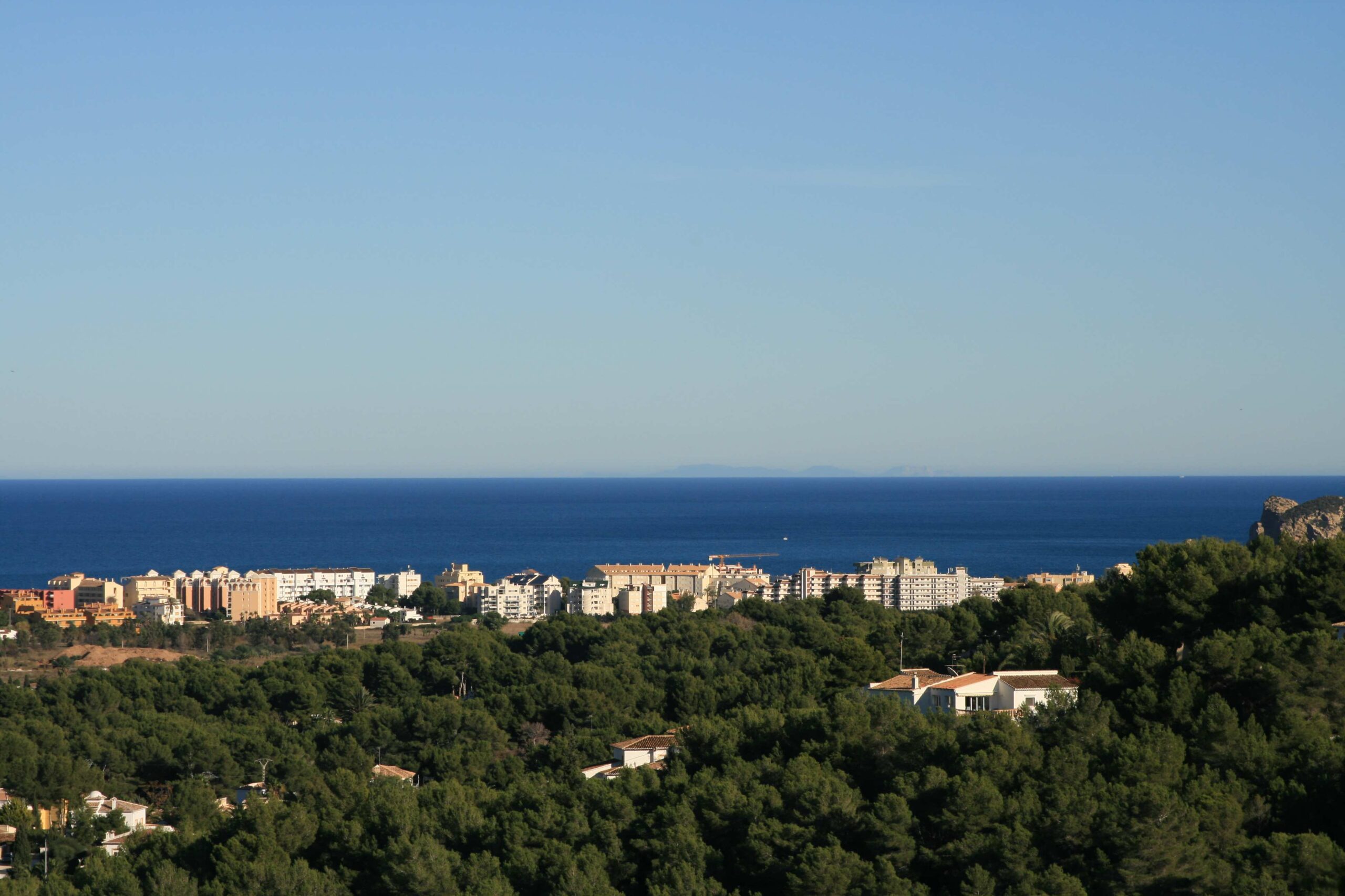 Vista panorámica desde la parcela que abarca un denso pinar en primer plano, el núcleo urbano costero y el mar Mediterráneo bajo un cielo despejado.