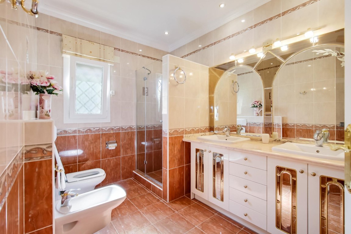 Baño completo con lavabo doble, espejos de arco con iluminación integrada, ducha de cristal y revestimiento cerámico en tonos cálidos.