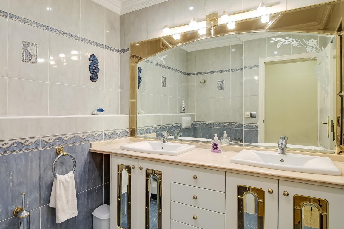 Baño principal con lavabo doble sobre encimera de mármol, mueble con detalles dorados, espejo de gran formato y revestimiento cerámico en tonos azules.