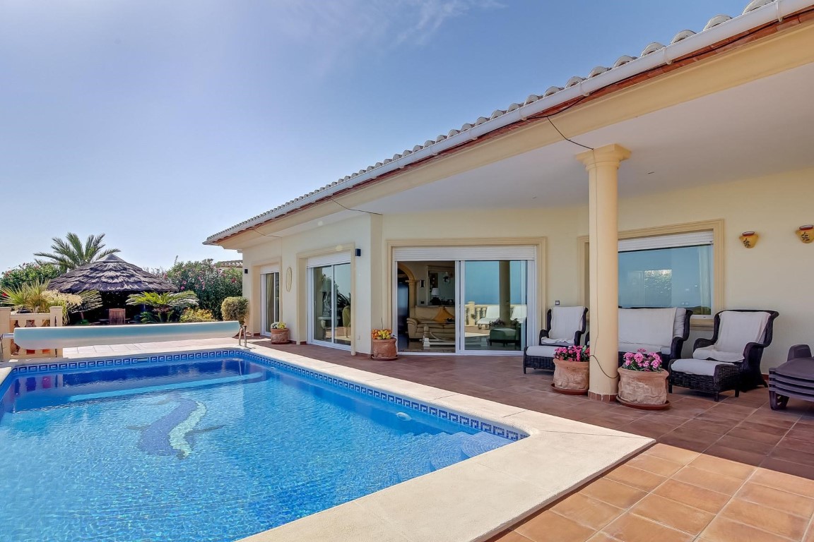 Terraza principal con piscina de mosaico, porche cubierto sostenido por columnas y zona de estar. Al fondo destaca un cenador tropical rodeado de vegetación mediterránea.