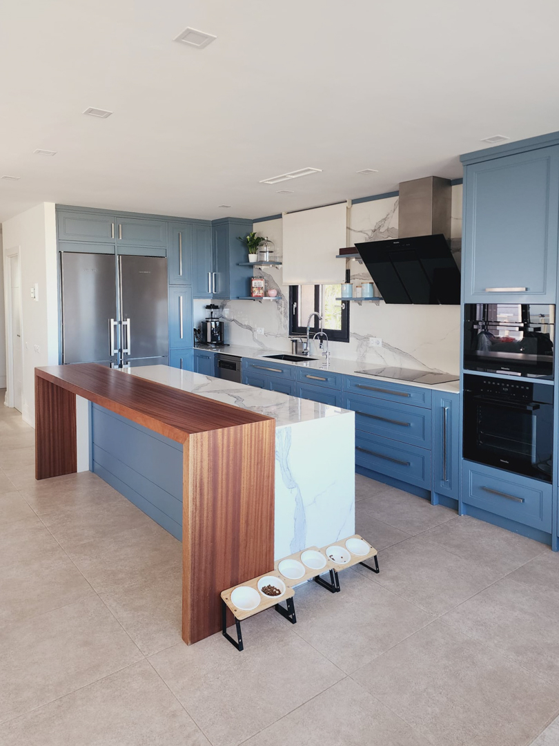 Cocina de diseño con mobiliario azul, isla central de mármol veteado y barra de madera integrada. Equipada con electrodomésticos Miele de alta gama y encimera a juego.