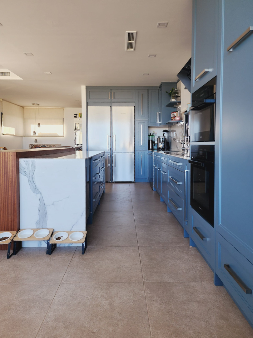 Cocina moderna con mobiliario azul pastel, isla central de mármol veteado y electrodomésticos Miele integrados. Suelo de gres porcelánico y aire acondicionado por conductos.