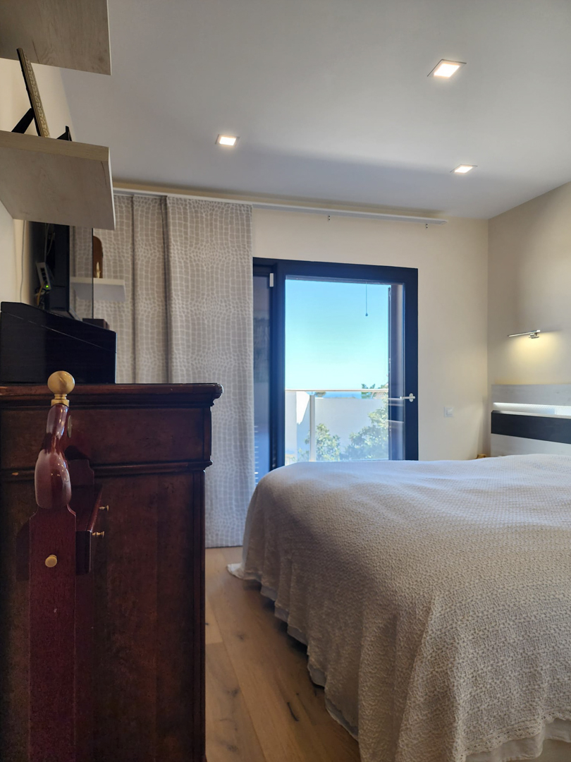 Dormitorio con suelos de madera, iluminación empotrada y acceso directo a la terraza con vistas al mar a través de un ventanal de marco oscuro.