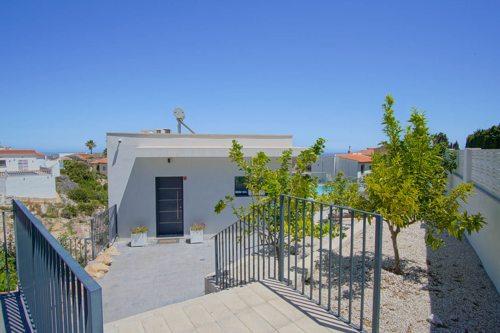 Entrada principal de la villa con puerta de seguridad gris, fachada blanca moderna, sistema solar térmico y jardín de grava con árboles frutales.