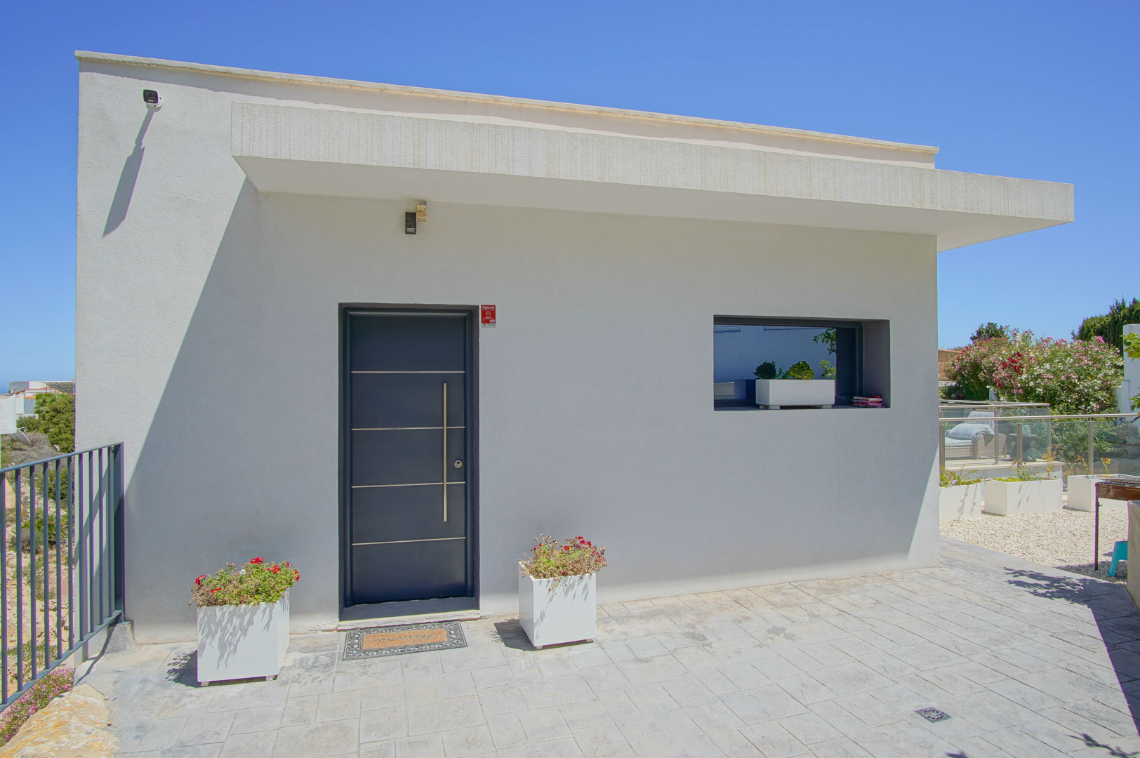 Fachada blanca minimalista con puerta de entrada gris oscuro de diseño moderno, tirador vertical de acero y maceteros decorativos en el acceso pavimentado.