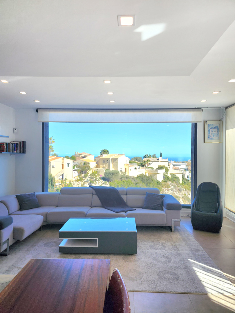 Salón luminoso con un gran ventanal panorámico hacia el mar, equipado con un sofá esquinero gris, mesa de centro moderna e iluminación LED empotrada.