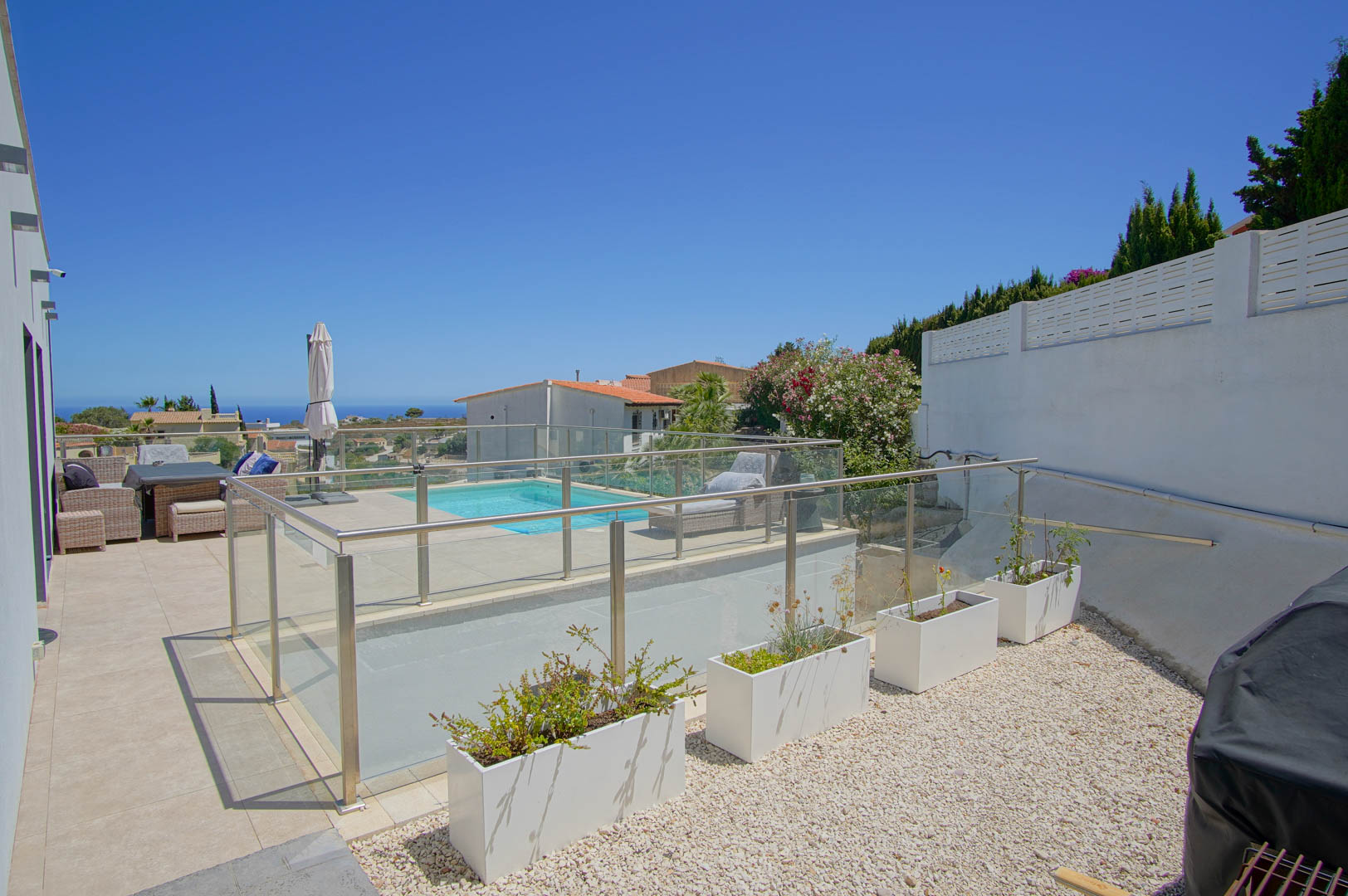 Terraza exterior con barandillas de cristal y acero, zona de piscina con solárium y área lateral de grava con jardineras blancas bajo un cielo despejado.