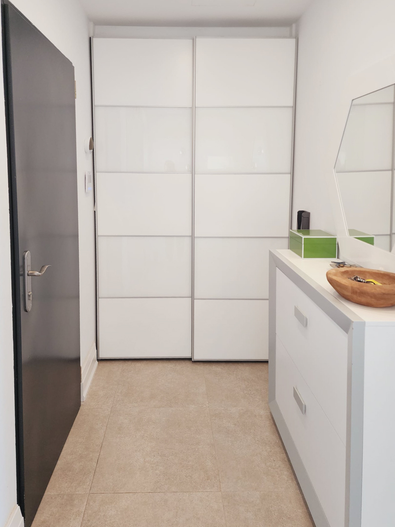 Vestíbulo de entrada con armario empotrado de puertas correderas blancas, cómoda minimalista y suelo de gres porcelánico en tonos arena.