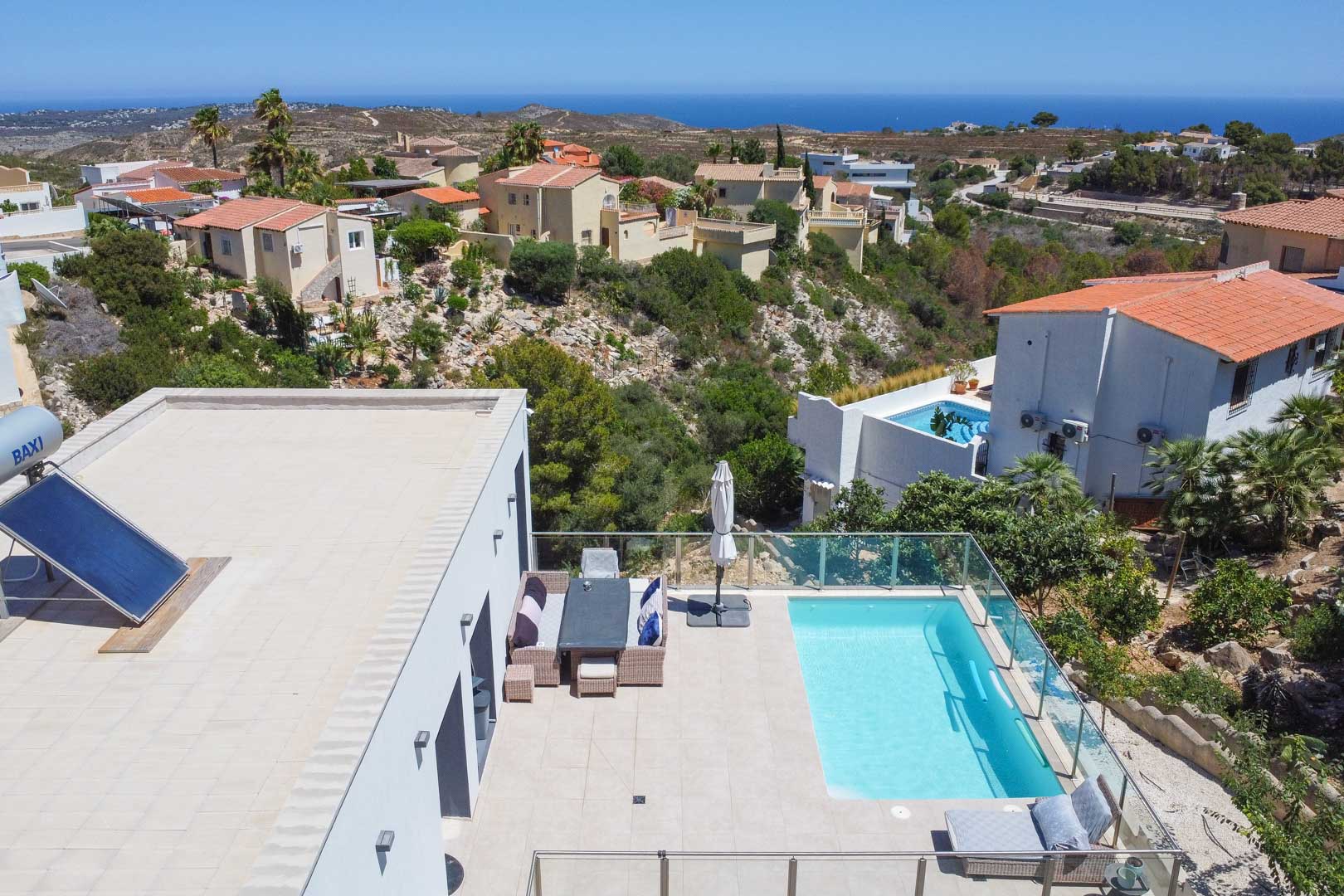 Vista aérea de la terraza con piscina rectangular, barandillas de cristal y comedor exterior. Incluye placa solar en cubierta y vistas panorámicas al Mediterráneo.