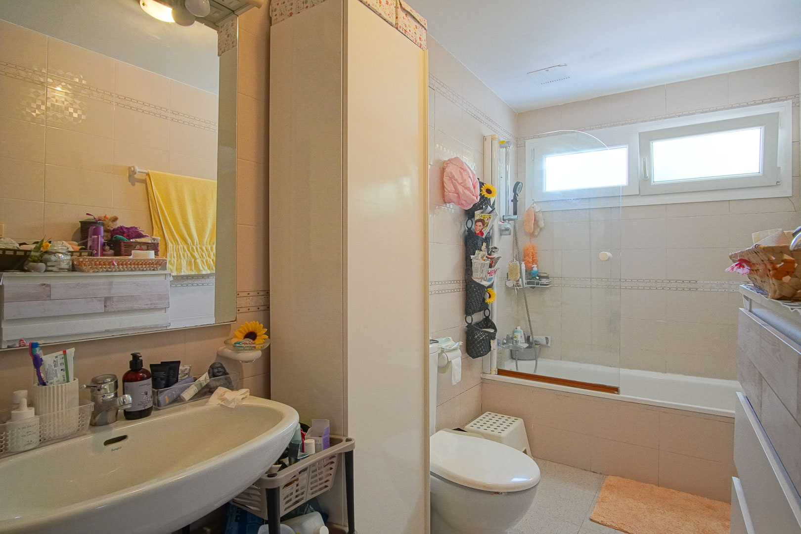 Baño completo con bañera y mampara de cristal, lavabo blanco y armario columna. Revestimiento cerámico en tonos crema y ventana para ventilación natural.